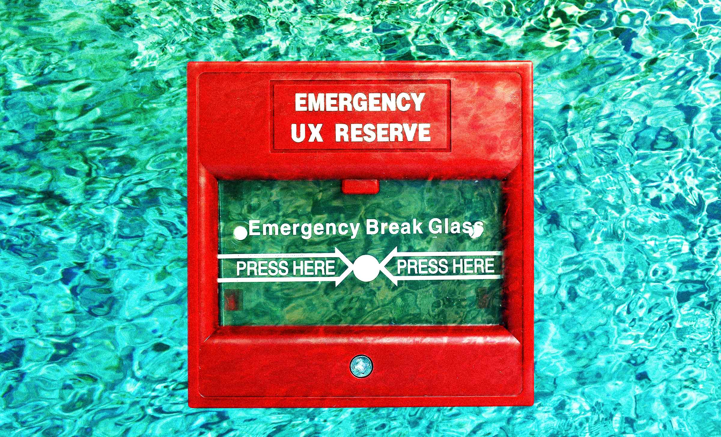 A red fire alarm behind a bright blue water background. The glass on the fire alarm says, "Emergency Break Glass," and printed on the alarm it reads, "Emergency: UX Reserve."