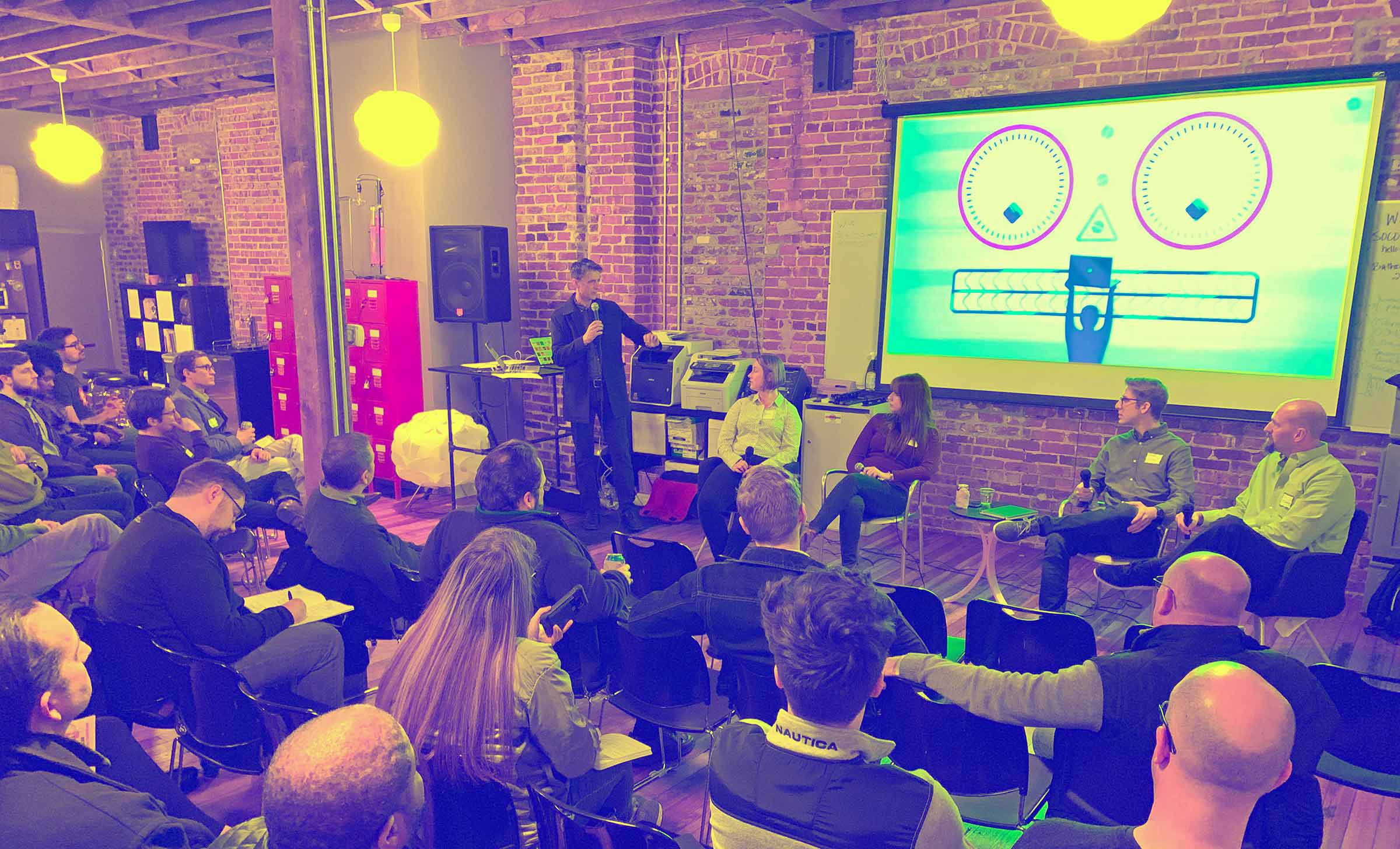 A panel, with the audience in chairs facing a speaker. Behind the speaker is a graphic of a face made out of computer parts.