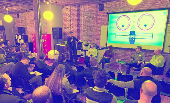 A panel, with the audience in chairs facing a speaker. Behind the speaker is a graphic of a face made out of computer parts.