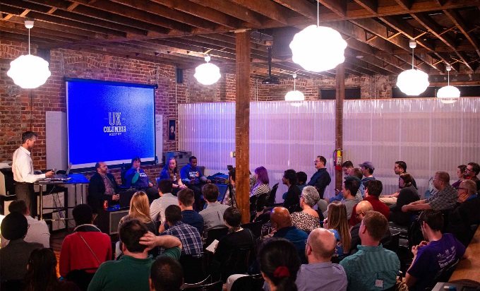 An audience of around thirty people sit looking towards a screen. In front of the screen stands a moderator and four panelists.