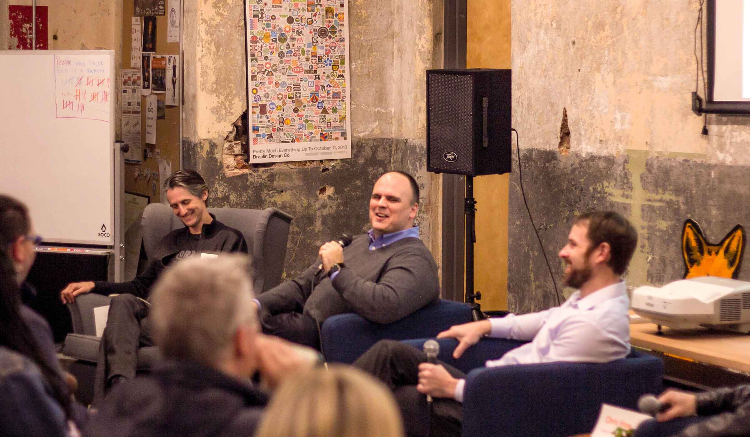 A panel of people talking to a crowd. The man in the middle holds a microphone.