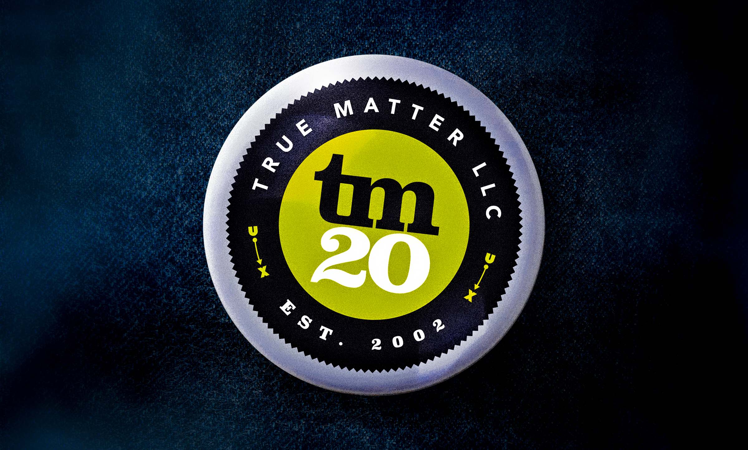 A silver, circular badge on a dark blue jean background, with a tm20 logo and text wrapped around it reading True Matter LLC, Est. 2002, embellished with a U connected to an X with an arrow.